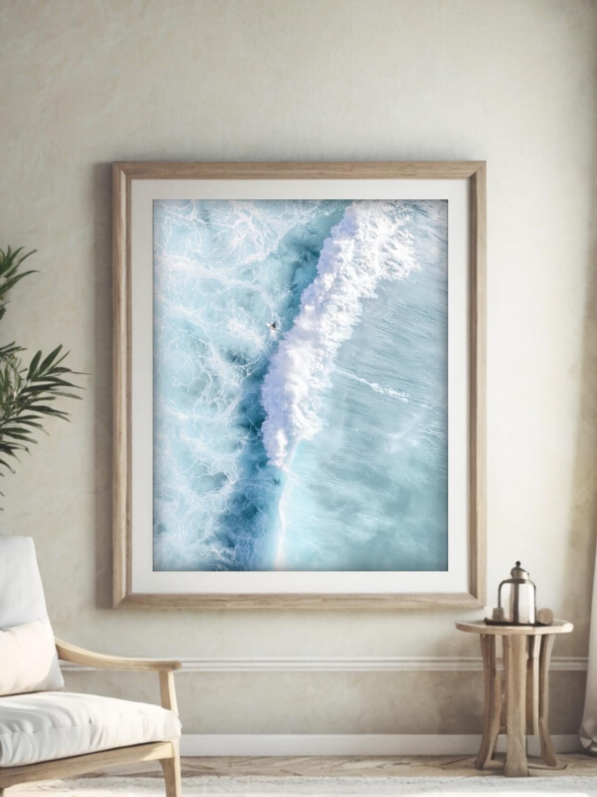 Framed ocean wave art print showcasing an aerial view of a surfer near a crashing wave, displayed on a white brick wall.