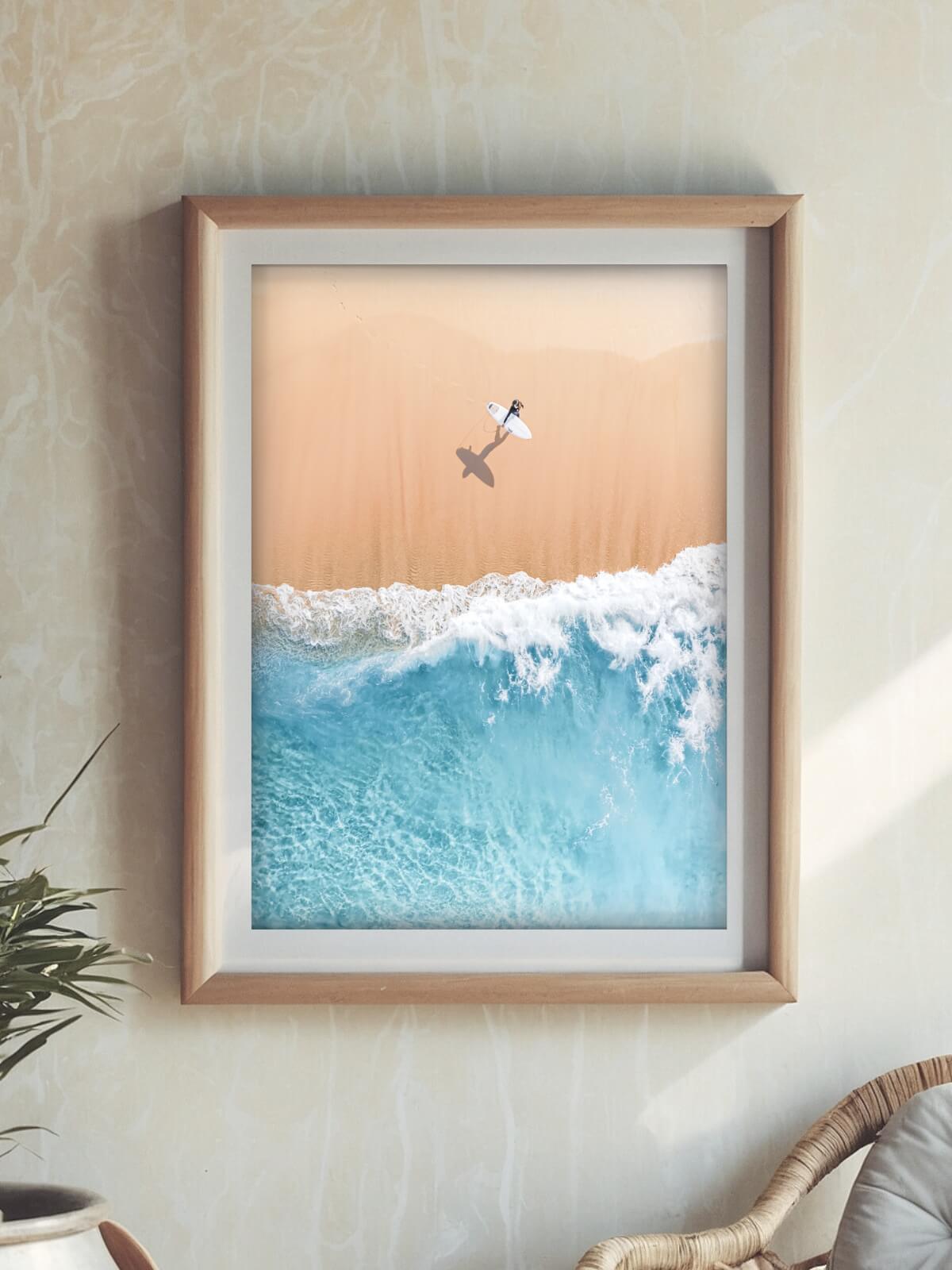 Framed aerial print of a surfer walking along sandy beach with waves, highlighting a serene coastal scene in vibrant blues and warm tones.