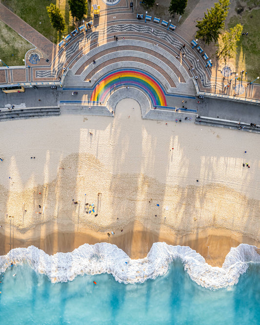 Coogee Pavilion with rainbow-painted steps and sandy beach below, captured from above on Sydney’s coastline.