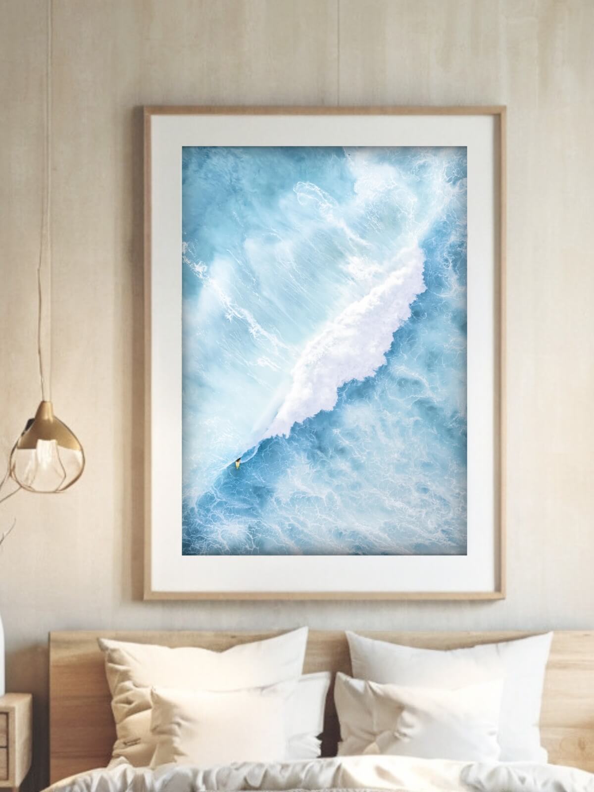 Framed ocean wave art print with a surfer displayed above a cozy bed in a minimalist bedroom.