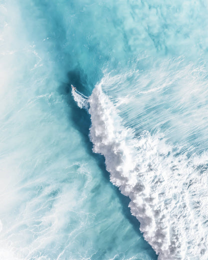 Aerial photo of a surfer riding along a foamy ocean wave with calm blue water.