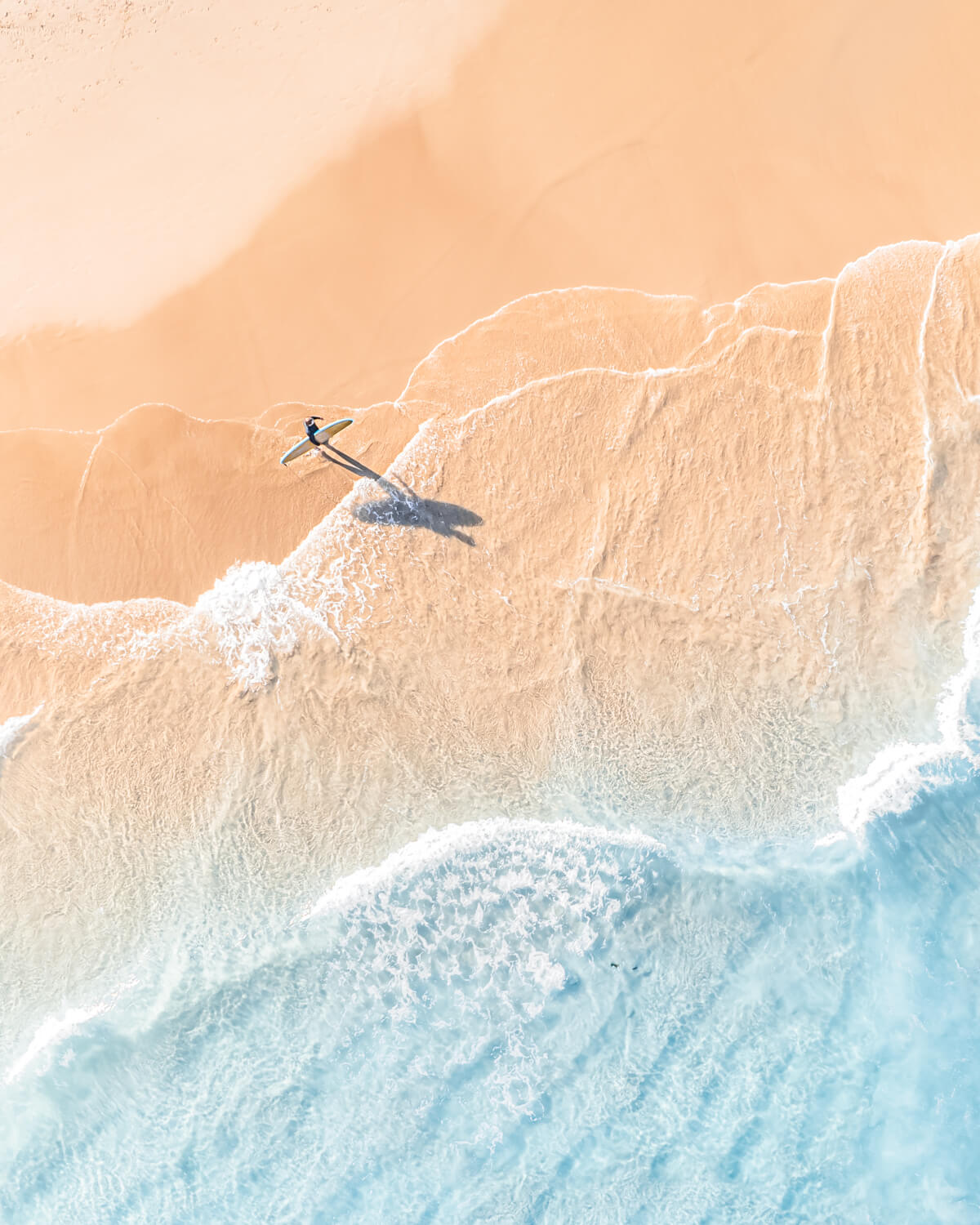 A lone surfer strolls back to the surf point on Byron Bay’s sunlit shore, casting a shadow over soft sands and tranquil waves.