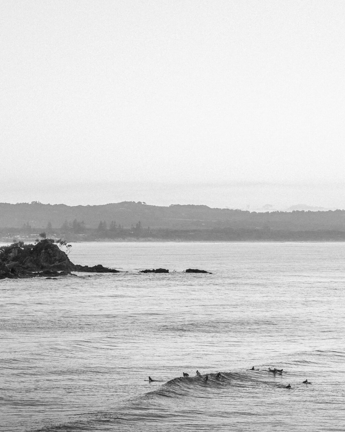 Black-and-white photo of surfers riding waves at Wategos Beach with Byron Bay and The Pass in the background.