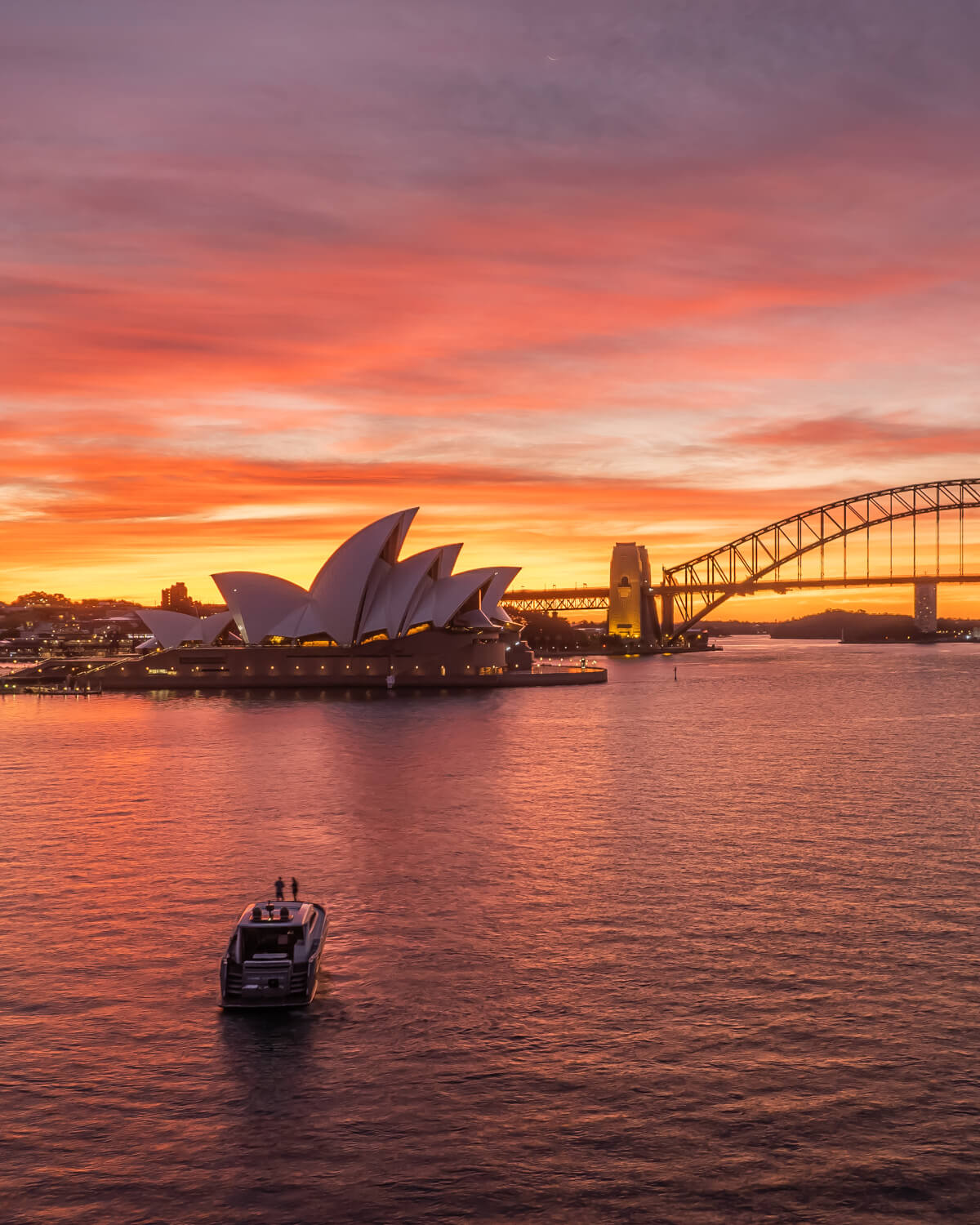 Photo of Sydney Harbour at sunset with orange skies, featuring the Opera House in intense lights