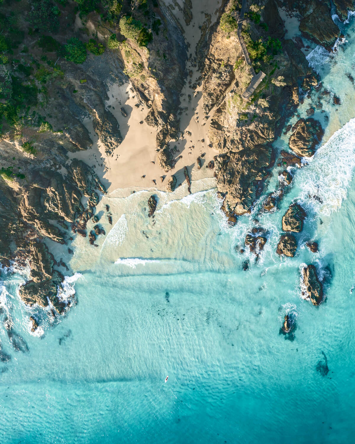 Aerial view of The Pass, Byron Bay, showcasing rocky cliffs and turquoise waters meeting sandy shore.