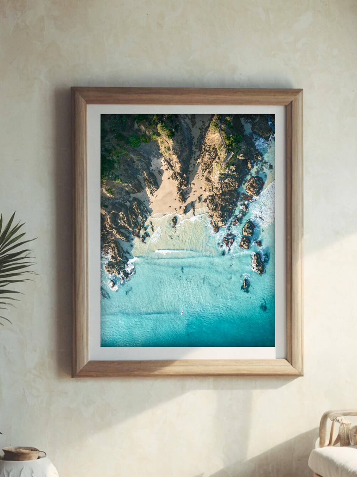 Framed aerial artwork of The Pass, Byron Bay, displaying rocky cliffs and turquoise waters meeting sandy shore.